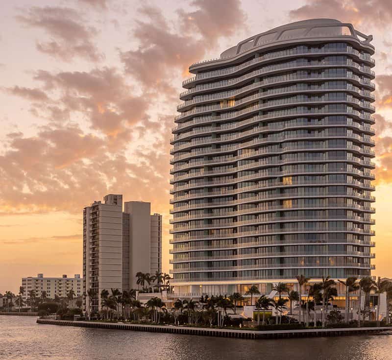 Modern waterfront skyscraper at sunset with palm trees.